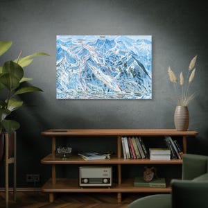 May include: A framed ski resort map featuring a detailed illustration of ski runs, lifts, and mountain terrain. The map is predominantly blue and white, with red lines indicating ski trails. The artwork is mounted on a wall above a wooden shelf.