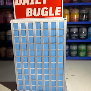 Peut inclure: Une maquette miniature du bâtiment Daily Bugle, avec une enseigne rouge et du texte blanc. Le bâtiment a une façade argentée avec de nombreuses fenêtres bleues. Le modèle est une représentation détaillée d'un gratte-ciel.