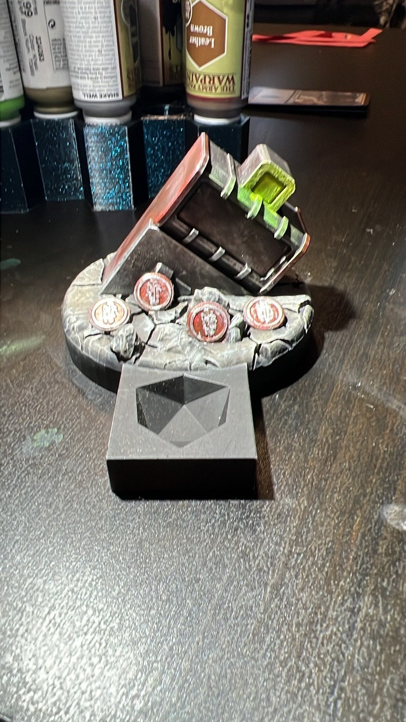 Puede incluir: Un modelo en miniatura de un cofre con una tapa verde brillante, sentado sobre una base de piedra gris con fichas rojas y blancas. El cofre est&aacute; sobre una base de espuma negra.