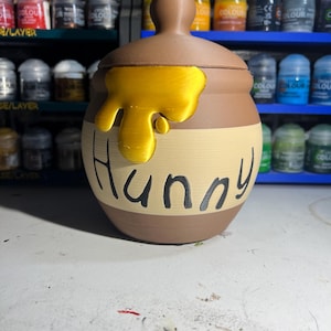 May include: A brown and tan decorative jar with a lid, featuring a gold-colored honey drip design. The word "Hunny" is written in black script across the front. The jar is likely a decorative item or container.