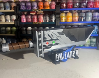 May include: A replica Buster Sword from Final Fantasy VII, featuring a silver and black blade with a brown handle. The sword has red and green accents and the Final Fantasy VII logo on the base.