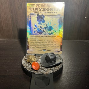 May include: A Magic: The Gathering card featuring Tinybones, the Pickpocket, a legendary creature with deathtouch. The card is a skeleton rogue with a gold and blue background. The card is displayed on a black stand with a cowboy hat and coins.