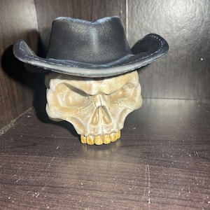 May include: A white skull figurine wearing a black cowboy hat. The skull has a detailed face with a prominent nose and a set of gold teeth.