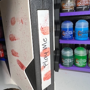 Puede incluir: Una caja blanca con una etiqueta negra que dice "Play Me" en letras rojas. La caja está cubierta de pintura roja salpicada. La caja está sobre una superficie de madera.