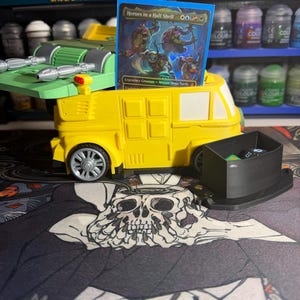 May include: A yellow toy van with green details, a card that reads "Heroes in a Half Shell" and a black container with dice. The card features the Teenage Mutant Ninja Turtles. Shelves of paint are in the background.
