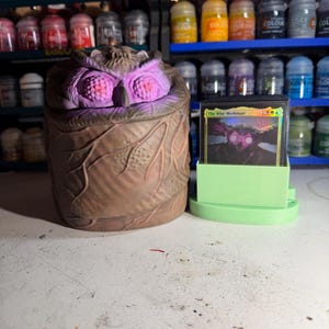 May include: A brown owl-shaped container with purple accents and a green card holder. The container has a textured surface and the text "The Wise Mothman" is visible on a card. Shelves of paint bottles are in the background.