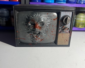 May include: A black and gray television with a bloody, distorted face on the screen. The television is a miniature model and has a red and white knob on the side.