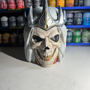 May include: A white skull with a silver crown and long white hair. The skull has a red and gold trim around the neck. The skull is a 3D printed model.