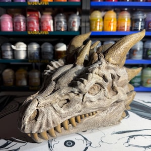 May include: A sculpted dragon head figurine featuring a weathered, stone-like texture in shades of beige and grey. The creature has prominent horns, detailed scales, and a row of sharp, pointed teeth visible along the jawline in a profile view.