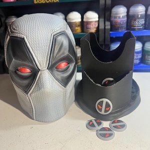 May include: A gray and black Deadpool mask with red eye lenses, a black stand, and three gray and red circular tokens. The mask and stand are on a white surface. Paint bottles are visible in the background.