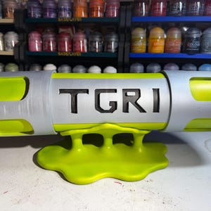 May include: A neon green and silver prop with the letters TGRI. The prop has a cylindrical shape with rounded ends and is supported by a matching green, blob-like base. The background features shelves of paint bottles.