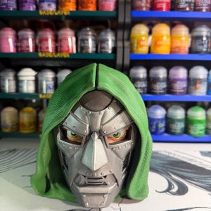 Op de afbeelding: Een gedetailleerde buste van een personage met een metallic zilveren masker met zichtbare klinknagels en een textuurrijke groene kap. Het masker heeft een scherp neusstuk, een gestructureerde kaaklijn en zichtbare groene ogen.