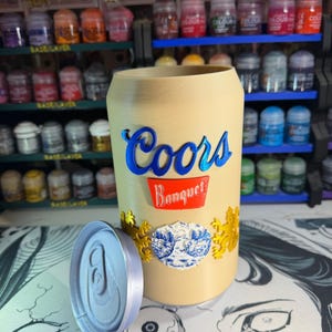 Peut inclure: Un récipient cylindrique beige en forme de canette de bière avec un couvercle amovible. L'extérieur présente le texte bleu Coors au-dessus d'une étiquette rouge avec le texte blanc Banquet, ainsi que des emblèmes de lions dorés et un motif montagne.