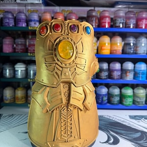 May include: A gold-colored Infinity Gauntlet replica featuring a large central yellow gem and five smaller colored gems in red, orange, purple, and blue. The gauntlet has a metallic finish with intricate mechanical detailing and molded plating.