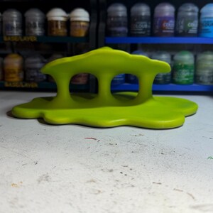 May include: A bright lime green, abstract object with three finger holes. The object has a flowing, organic design and sits on a white surface. The background features shelves filled with small paint jars.