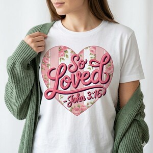 May include: White t-shirt featuring a pink heart design with the words "So Loved" in a cursive font. The heart is decorated with pink roses and vertical stripes, and the text "John 3:16" is below the heart.
