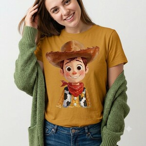 May include: A mustard yellow short-sleeved t-shirt featuring a graphic illustration of a cartoon cowboy character wearing a brown hat, a red bandana, and a cow-print vest. The character has large brown eyes and a friendly smile on its face.