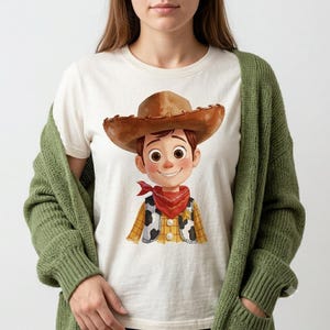 May include: An off-white t-shirt featuring a cartoon illustration of a character wearing a brown cowboy hat, a red bandana, and a yellow and white cow-print vest. The character has brown hair, large eyes, and a smiling expression, layered under a green cardigan.