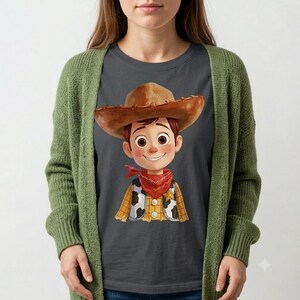 May include: A dark gray t-shirt featuring a cartoon illustration of a character wearing a brown cowboy hat, a red bandana, and a yellow shirt with a black and white cow-print vest, layered under a long-sleeved olive green open-front knit cardigan.