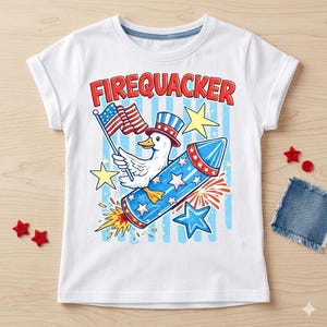 May include: White t-shirt with a colorful graphic featuring a duck wearing a patriotic hat and holding an American flag, riding a blue rocket with stars. The word "FIREQUACKER" is printed in red above the graphic.