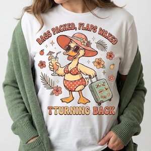 May include: White t-shirt featuring a cartoon duck in a swimsuit, sunglasses, and a sun hat, holding a cocktail and a suitcase. The shirt has the text "BAGS PACKED, FLAPS WAXED, TURNING BACK" in a retro font, with floral and tropical accents.