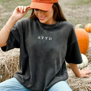 May include: An orange corduroy baseball cap with the text "PUMPKIN SPICE" is worn with a dark gray t-shirt with the letters "ATYD". The person is sitting on hay bales with pumpkins in the background.
