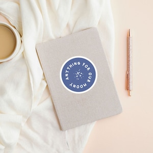 May include: A blue and white sticker with the text "Anything for our moony" and stars inside a circle, on a beige notebook.