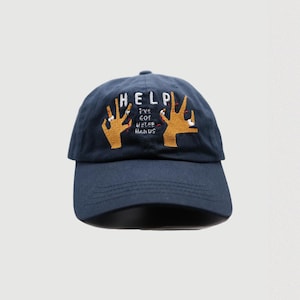 May include: A navy blue baseball cap with the word "HELP" embroidered in gold on the front. Two yellow hands with red fingernails are embroidered below the word "HELP" with the text "I'VE GOT HELPER HANDS" in white.