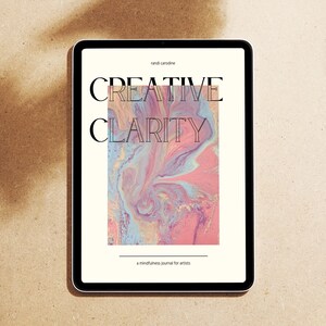 May include: A tablet computer displaying a book cover with the title "Creative Clarity: A Mindfulness Journal for Artists" in black text on a white background. The cover features a pink, blue, and yellow abstract marbled design.