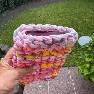 Könnte beinhalten: Handgefertigte Tablet-Hülle in Rosa, Gelb und Lila. Die Hülle besteht aus dickem, geflochtenem Stoff und trägt ein Etikett mit der Aufschrift "Hake...by". Das Tablet ist teilweise in der Hülle sichtbar.