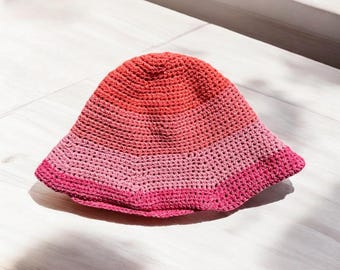 May include: A pink and red crocheted bucket hat. The hat has a wide brim and is made of a soft, textured yarn.