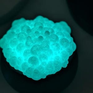 May include: A glowing, luminous turquoise object with a cluster of numerous small, rounded, bubble-like protrusions covering its surface. The object emits a bright, vibrant light, creating a high-contrast effect against the dark surroundings.