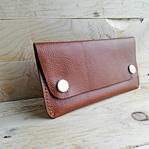 May include: A brown leather wallet with two gold metal snaps. The wallet is lying on a light wood surface.
