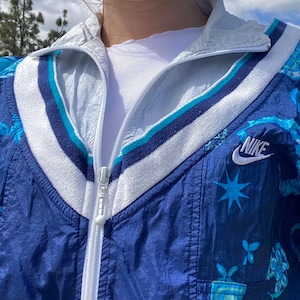 May include: A blue and white Nike track jacket with a floral pattern and a white zipper. The jacket has a white collar and stripes in shades of blue. The Nike logo is visible on the right side of the jacket.