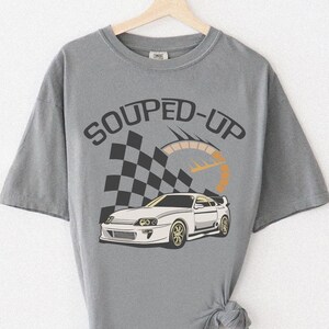 May include: Gray t-shirt with a graphic of a white sports car, a checkered flag, and the words "SOUPED-UP". The design includes a speedometer graphic. The shirt is hanging on a wooden hanger against a white wall.