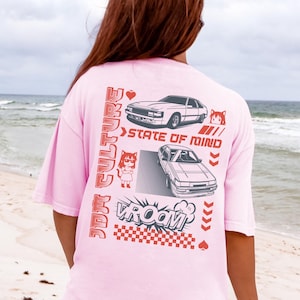 May include: A light pink t-shirt with a graphic design featuring two cars, anime-style characters, and the words "JDM Culture" and "State of Mind" in red. The shirt has a relaxed fit and short sleeves.