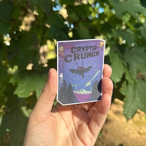 May include: A sticker of a cereal box with the text "Cryptid Crunch" and a cartoon image of a moth, a ghost, and a plesiosaur eating the cereal.