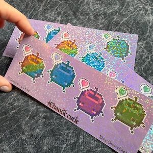 May include: A rectangular holographic sticker sheet featuring pixel art characters in various colors, each with a small heart speech bubble. The text "Chaotic Cutie" is printed in the center, with "Chaotic-Cutie" and "Etsy: ChaoticCutieArt" text.