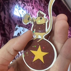 May include: A sticker featuring a cartoon mouse in a band uniform playing a trumpet. The mouse stands on a brown ball with a yellow star. The sticker has a white border and holographic accents.
