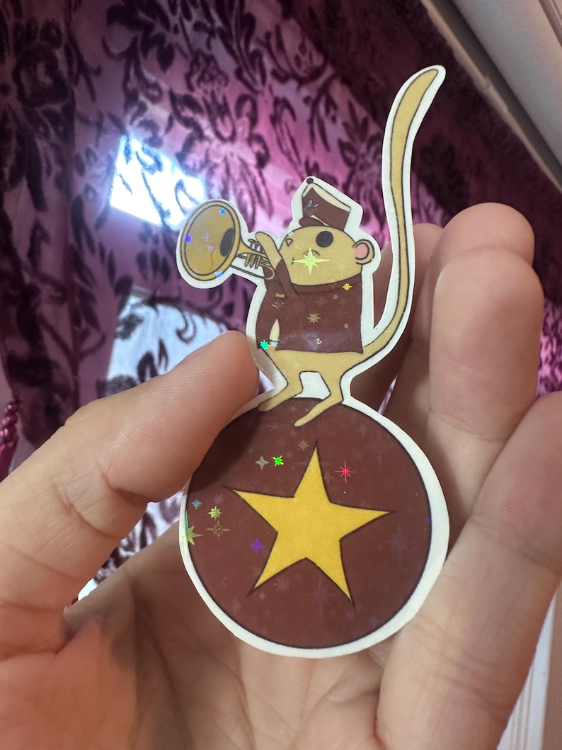 May include: A sticker featuring a cartoon mouse in a band uniform playing a trumpet. The mouse stands on a brown ball with a yellow star. The sticker has a white border and holographic accents.