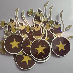 May include: A collection of stickers featuring cartoon monkeys playing trumpets atop brown orbs with gold stars. The monkeys wear brown jackets and hats. The orbs have a sparkly, iridescent effect.