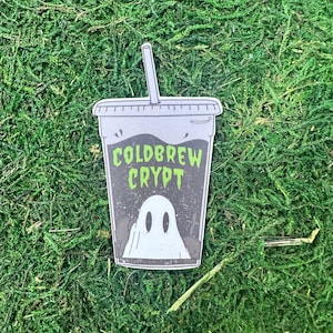 May include: A black and white illustration of a ghost in a coffee cup with a straw. The cup says "Coldbrew Crypt".