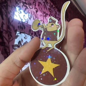 May include: A sticker featuring a cartoon monkey in a band uniform playing a trumpet. The monkey stands on a brown ball with a yellow star. The sticker has a holographic effect with colorful star accents.