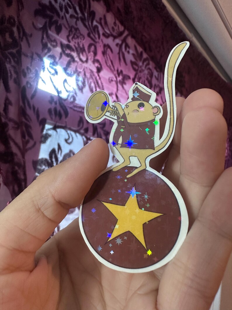 May include: A sticker featuring a cartoon monkey in a band uniform playing a trumpet. The monkey stands on a brown ball with a yellow star. The sticker has a holographic effect with colorful star accents.