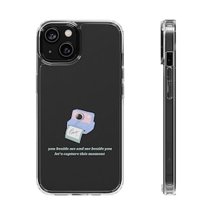 Peut inclure: Une coque de téléphone transparente avec un fond noir et une illustration de dessin animé d'un appareil photo Polaroid rose et bleu avec une photo qui dit "love" qui en sort. Le texte "you beside me and me beside you let's capture this moment" est imprimé sous l'image.