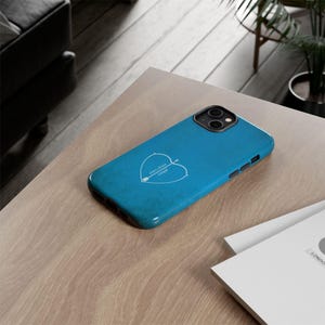 May include: A teal phone case with a white heart and arrow design. The case has a glossy finish and is lying on a light wood surface. The text "arian share" and "angel" is inside the heart.