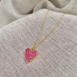 May include: A gold chain necklace with a pink heart pendant. The heart is filled with a textured pink material and has a gold border.