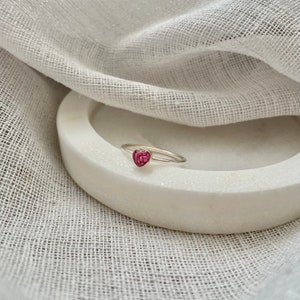 Peut inclure: Une bague en argent avec une pierre en forme de c&oelig;ur rose. La bague est sur une surface blanche.