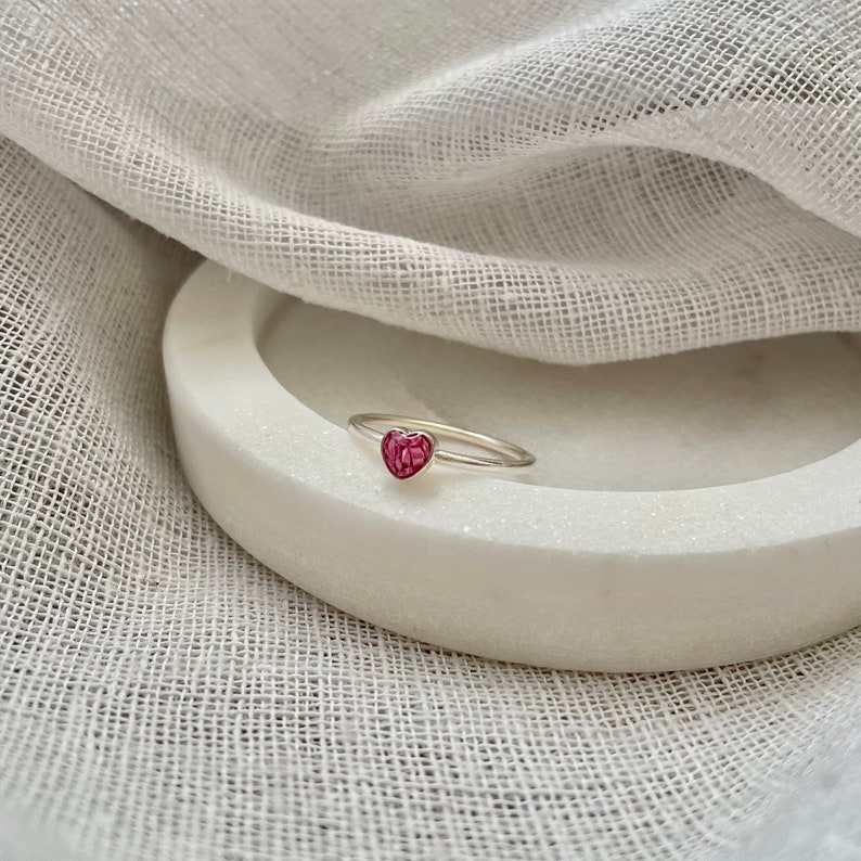 Peut inclure: Une bague en argent avec une pierre en forme de c&oelig;ur rose. La bague est sur une surface blanche.
