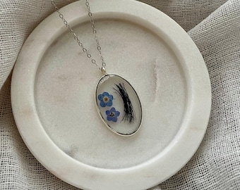 May include: A silver chain necklace with an oval pendant featuring two blue forget-me-not flowers and a black eyelash encased in clear resin.